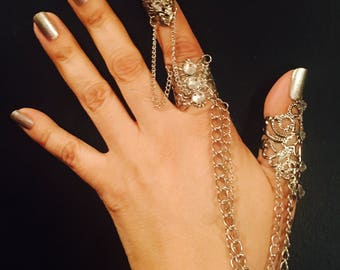 May include: Silver-toned finger rings connected by chains. The rings feature ornate filigree designs and clear gemstones. One ring covers the entire finger, while others adorn the knuckles. The jewelry is displayed against a dark background.