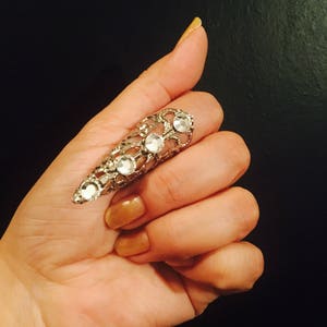 Puede incluir: Un anillo de plata con diamantes de imitación transparentes en forma de garra. El anillo se lleva en el dedo índice.