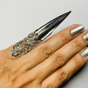 May include: Silver-colored nail ring with a long, pointed tip. The ring features an ornate, filigree design on the finger band. The nail ring is worn on a finger with silver nail polish.