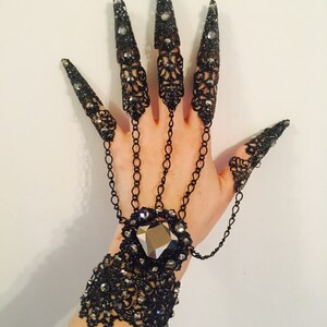 K&ouml;nnte beinhalten: Schwarzer Filigran-Handschmuck mit langen, spitzen Nagelbedeckungen. Das Accessoire im Gothic-Stil verf&uuml;gt &uuml;ber ein Armband mit einem gro&szlig;en, facettierten Edelstein und Kettendetails, die mit den Nagelbedeckungen verbunden sind. Die Nagelbedeckungen sind mit kleinen Kristallen verziert.