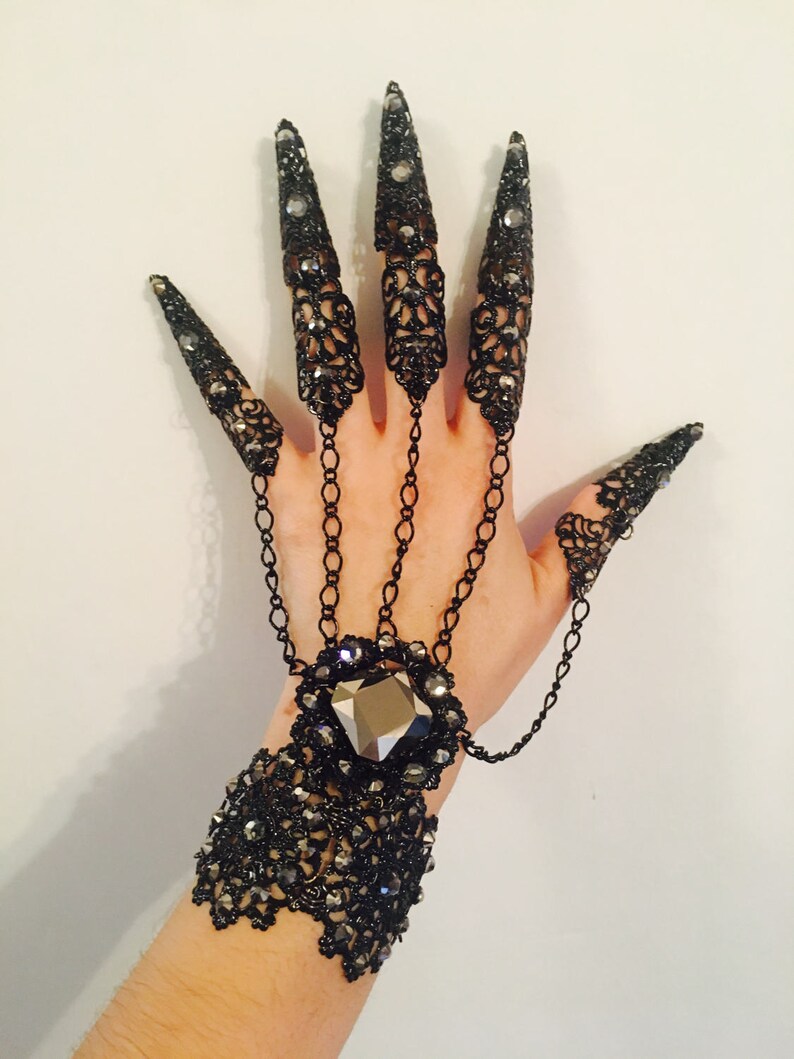 K&ouml;nnte beinhalten: Schwarzer Filigran-Handschmuck mit langen, spitzen Nagelbedeckungen. Das Accessoire im Gothic-Stil verf&uuml;gt &uuml;ber ein Armband mit einem gro&szlig;en, facettierten Edelstein und Kettendetails, die mit den Nagelbedeckungen verbunden sind. Die Nagelbedeckungen sind mit kleinen Kristallen verziert.