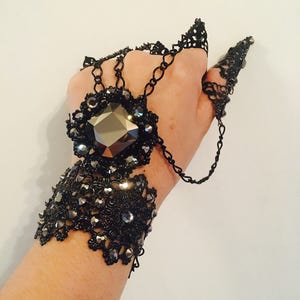 K&ouml;nnte beinhalten: Schwarzer Filigran-Handschmuck mit einem gro&szlig;en, facettierten, grauen Edelstein in der Mitte. Das aufwendige Design umfasst Ketten und spitze Fingerakzente, die eine gotische oder Fantasy-&Auml;sthetik erzeugen. Das armbandartige St&uuml;ck ist mit kleineren, funkelnden Kristallen verziert.