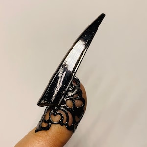 May include: Black nail armor ring with a long, pointed tip. The ring features an ornate, openwork design around the finger base. The nail armor is a dark, glossy black, creating a striking contrast against the skin.