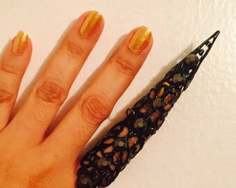 May include: A black, ornate finger ring with a long, pointed design. The ring is embellished with small, dark-colored stones and intricate metalwork. The nails are painted gold.