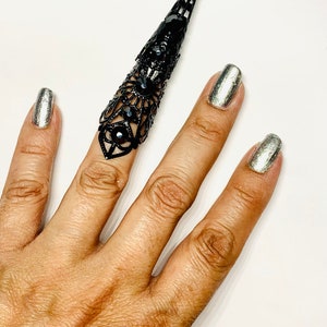 May include: A black metal ring with a pointed design and black rhinestones. The ring is worn on the index finger of a hand.