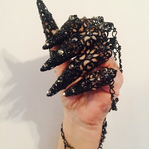 K&ouml;nnte beinhalten: Schwarze Spitzen-Fingerpanzerung mit langen, spitzen Spitzen, verziert mit kleinen, funkelnden Verzierungen. Das aufwendige Design verf&uuml;gt &uuml;ber eine zarte Kette, die sich um das Handgelenk wickelt und ein gotisches oder Fantasy-inspiriertes Accessoire schafft.