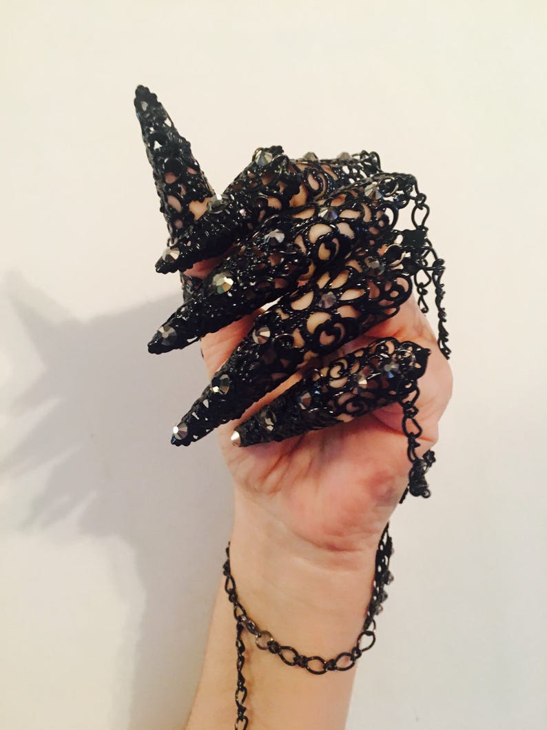 K&ouml;nnte beinhalten: Schwarze Spitzen-Fingerpanzerung mit langen, spitzen Spitzen, verziert mit kleinen, funkelnden Verzierungen. Das aufwendige Design verf&uuml;gt &uuml;ber eine zarte Kette, die sich um das Handgelenk wickelt und ein gotisches oder Fantasy-inspiriertes Accessoire schafft.