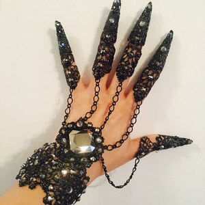 K&ouml;nnte beinhalten: Schwarzer Metallhandschmuck mit langen, spitzen Nagelbedeckungen, die durch Ketten mit einem Armband verbunden sind. Das Armband ist mit Strasssteinen und einem gro&szlig;en, facettierten Mittelstein verziert. Der Schmuck hat eine gotische oder Fantasy-&Auml;sthetik.