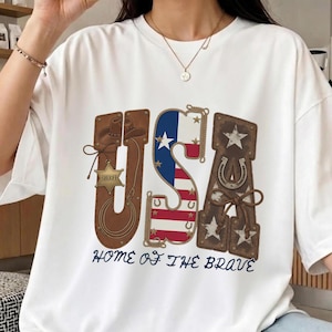 Peut inclure: T-shirt blanc avec un motif patriotique USA. Le motif présente les lettres "USA" dans une police de style western, incorporant des éléments comme un chapeau de cowboy, le drapeau américain et un fer à cheval. Le texte "HOME OF THE BRAVE" est en dessous.