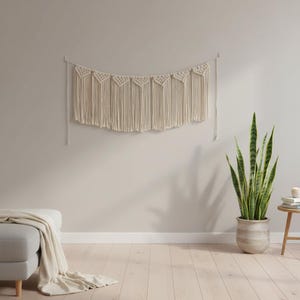 May include: A cream-colored macrame wall hanging with a geometric design. The wall decor is suspended from a wooden dowel and features a series of triangular patterns and long fringe. The neutral tones complement the light-colored wall.