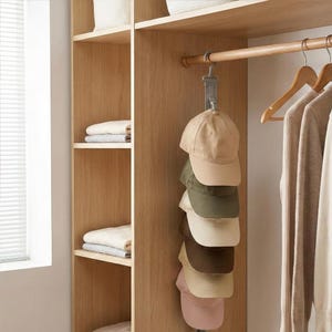 Puede incluir: Un armario de madera con estantes y una barra para colgar. Un organizador de sombreros gris sostiene seis gorras de béisbol en varios colores: beige, verde oliva, crema, marrón y rosa. Perchas de madera sostienen suéteres de colores neutros.