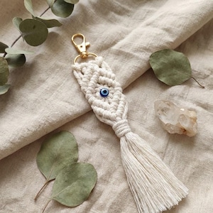 May include: Cream-colored macrame keychain with a gold-tone clasp and a blue evil eye bead. The keychain features a woven design with a tassel at the bottom. The background includes eucalyptus leaves and a neutral-toned fabric.