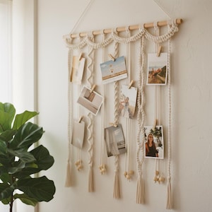 May include: A macrame photo display with a wooden dowel and hanging cords. Small wooden clothespins hold photos. The design features knotted details, tassels, and wooden beads. The color palette is neutral, with off-white and natural wood tones.