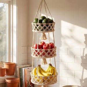 Puede incluir: Una cesta colgante de macramé de tres niveles llena de productos frescos. La cesta superior contiene aguacates verdes, la del medio manzanas rojas y la inferior plátanos amarillos. Las cestas están suspendidas de una viga de madera.