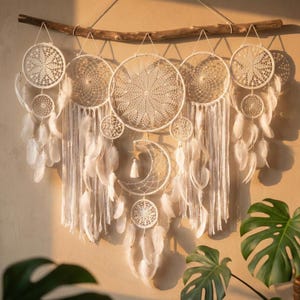 Könnte beinhalten: Ein weißer Traumfänger-Wandbehang mit mehreren kreisförmigen und halbmondförmigen Designs. Der Traumfänger hat komplizierte Spitzmuster, weiße Federn und einen Ast zum Aufhängen. Das Design ist im Bohemian-Stil gehalten.