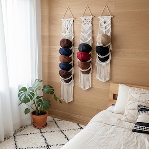 May include: Three macrame hat organisers hanging on a wooden wall, each holding several baseball caps. The caps are various colours, including brown, navy, red, and black. A potted plant and a bed with pillows are in the foreground.
