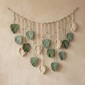 May include: A macramé wall hanging with leaf-shaped designs in green and cream. The leaves are suspended from a wooden dowel with wooden beads, creating a decorative and natural aesthetic. This piece is perfect for adding a touch of bohemian style to any space.