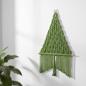 May include: A green macrame wall hanging in a triangular shape, resembling a Christmas tree. The handmade piece features intricate knotting patterns and a fringe at the bottom. A wooden dowel and a small wooden ring are used for hanging.
