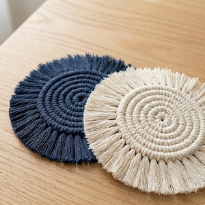 Pode incluir: Duas bases para copos redondas em macramé. Uma é azul marinho, a outra é branco sujo. Ambas têm um design em espiral no centro e franjas nas bordas. Estão colocadas sobre uma superfície de madeira castanha clara.