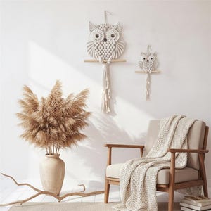 Könnte beinhalten: Zwei Wandbehänge in Form von Eulen aus Makramee, eine große und eine kleine, werden an einer weißen Wand gezeigt. Die Eulen sind aus cremefarbenem Seil gefertigt und sitzen auf Holzstäben. Eine Vase mit getrocknetem Pampasgras und ein Holzstuhl mit einer cremefarbenen Decke vervollständigen die Szene.
