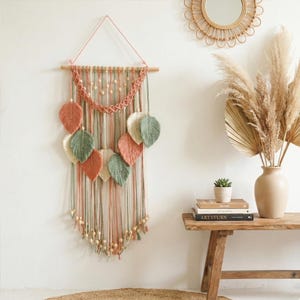 Op de afbeelding: Een macramé wandkleed met een houten stok, een geweven bovendeel en hangende strengen. Het ontwerp bevat bladvormige accenten in perzik-, groen- en crèmetinten, met houten kralen en kwasten. Wanddecoratie in bohemien stijl.