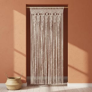 May include: Cream macrame curtain hanging in a doorway. The curtain has a detailed woven top with diamond and geometric patterns, and long, vertical strands. A woven basket sits on the floor in front of a peach-coloured wall.