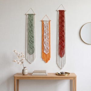 May include: Three macrame wall hangings in shades of green, orange, and red, each featuring a leaf design. The hangings are suspended from wooden dowels and triangular hangers. The wall decor is displayed on a white wall above a wooden console table.