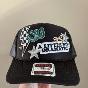 May include: Black trucker hat with a mesh back and a curved brim. The front features patches including a lightning bolt, stars, and the phrase "A Little Bit Dramatic". The hat has a snap closure and a "OTTO" label.
