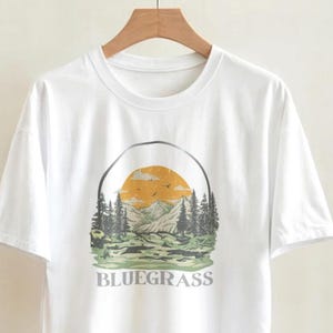 May include: A white short-sleeved t-shirt featuring a graphic print of a mountain landscape with pine trees, a yellow sun, and birds. The word BLUEGRASS is printed in a serif font below the illustration. The shirt is displayed on a wooden hanger.