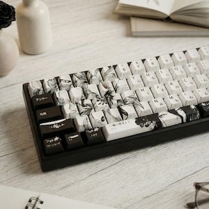 May include: A black and white mechanical keyboard with custom keycaps. The keycaps feature a floral design and Japanese characters. The keyboard is on a light-colored wooden surface.