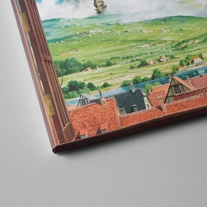 May include: A framed canvas print depicting a European village scene. The artwork features red-tiled roofs, green fields, a river, and a cloudy sky. The canvas is framed in a dark brown wooden frame.