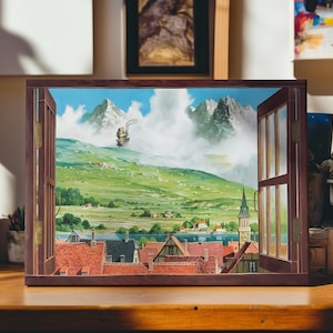 May include: A framed artwork depicting a landscape scene viewed through an open window. The artwork features a green hillside, a town with red-tiled roofs, and a mountain range in the background. The window frame is brown, and the sky is blue with clouds.