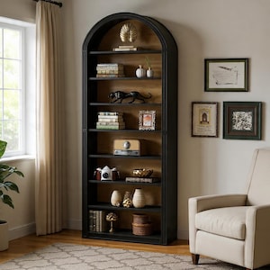 May include: A tall, black arched bookcase filled with decorative items. The shelves display books, vases, a panther figurine, a vintage radio, and framed pictures. The bookcase is set against a light-colored wall with a window and a beige armchair.