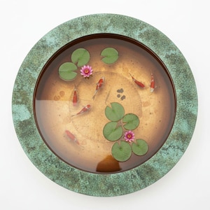 Puede incluir: Un elemento acuático redondo de color bronce con un borde verde desgastado. El agua contiene varios peces koi, nenúfares y nenúfares rosas. El elemento acuático está sobre un fondo blanco.