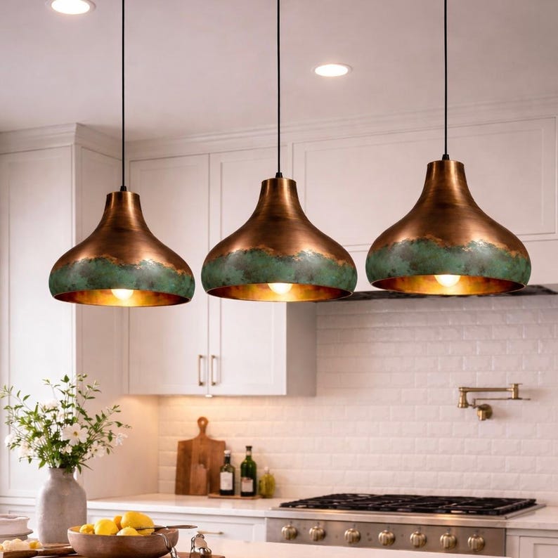 May include: Three pendant lights with a copper finish and a green patina band. The lights hang from black cords and illuminate a kitchen. The lights have a bell shape and a warm, inviting glow.