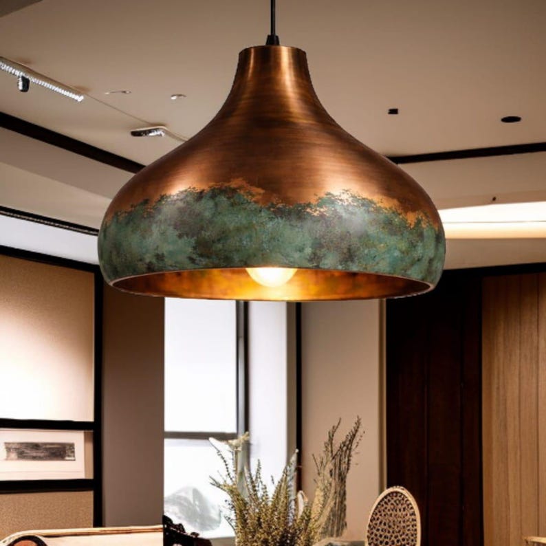 May include: A copper-colored pendant light fixture with a wide, rounded shade. The shade has a green patina band near the bottom, and a warm light shines from within. The fixture hangs from a black cord, suitable for interior lighting.