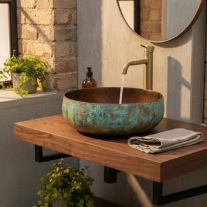 Puede incluir: Un lavabo de cobre con acabado verdoso se encuentra sobre un tocador de madera oscura. Un grifo dorado vierte agua en el lavabo. También se ven un espejo redondo y una toalla pequeña. El baño tiene una estética moderna y rústica.
