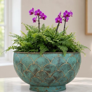 May include: A large, teal-colored planter with a scalloped design and gold accents, filled with green ferns and vibrant purple orchids. The planter sits on a white marble surface, creating a striking contrast. A decorative piece for indoor or outdoor use.