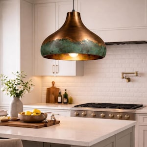 May include: A copper-colored pendant light with a green patina around the bottom edge hangs above a kitchen island. The light fixture has a rounded, bell-like shape and a single exposed bulb. The kitchen features white cabinets and a white countertop.