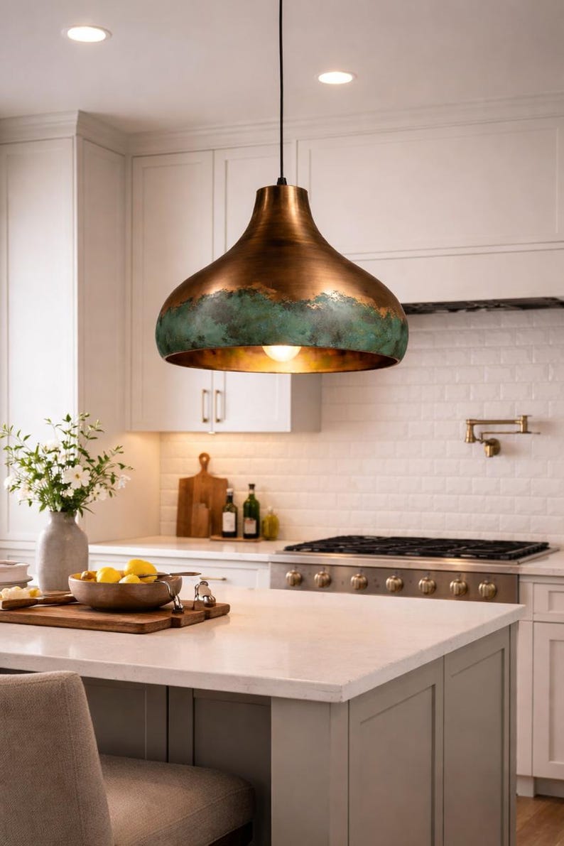May include: A copper-colored pendant light with a green patina around the bottom edge hangs above a kitchen island. The light fixture has a rounded, bell-like shape and a single exposed bulb. The kitchen features white cabinets and a white countertop.