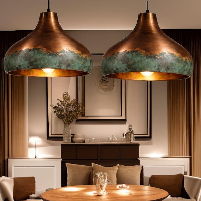 May include: Two copper-colored pendant lights with a verdigris band around the bottom edge. The lights have a bell shape and hang from black cords. The lights are illuminated, casting a warm glow.