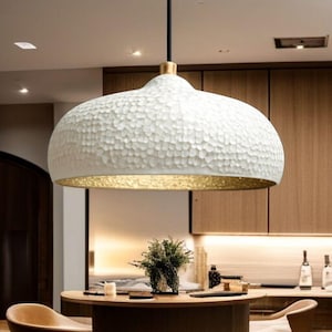 Könnte beinhalten: Eine weiße, strukturierte Pendelleuchte mit goldfarbenem Innenraum hängt von der Decke. Die Leuchte hat eine abgerundete, glockenförmige Form und wird von einem schwarzen Kabel abgehängt. Der Hintergrund zeigt Holzschränke und einen runden Tisch.