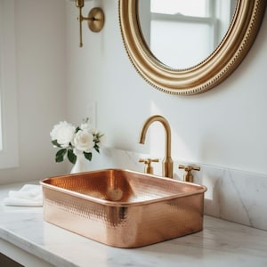 May include: A rectangular copper sink with a hammered texture sits on a marble countertop. A gold faucet and matching fixtures are installed. A gold-framed oval mirror hangs above the sink, reflecting the natural light from a nearby window.
