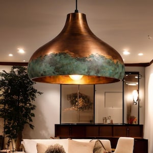 May include: A large, copper-colored pendant light fixture with a unique, inverted bell shape. The fixture has a distressed, verdigris green band around the middle, adding a touch of contrast. The interior is a warm gold color, and the light is on.