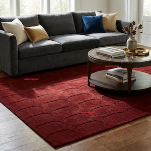 Puede incluir: Un salón con un sofá gris oscuro con cojines crema, dorados y azul marino. Una mesa de centro redonda está frente al sofá, y una alfombra roja oscura con un patrón ovalado cubre el suelo.