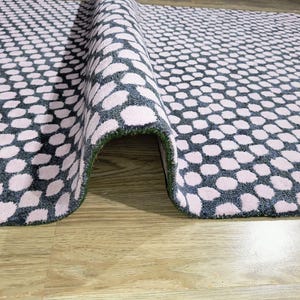 Op de afbeelding: Opgerolde vloerkleed met een herhalend patroon van roze ovalen op een donkergrijze achtergrond. De rand van het kleed toont een groene onderkant, op een licht houten oppervlak.