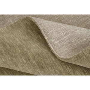 May include: A taupe-colored area rug with a dense, short-pile texture. The rug is folded over to reveal the underside, which displays a lighter, woven fabric construction with a visible grid-like pattern and a finished, reinforced edge.