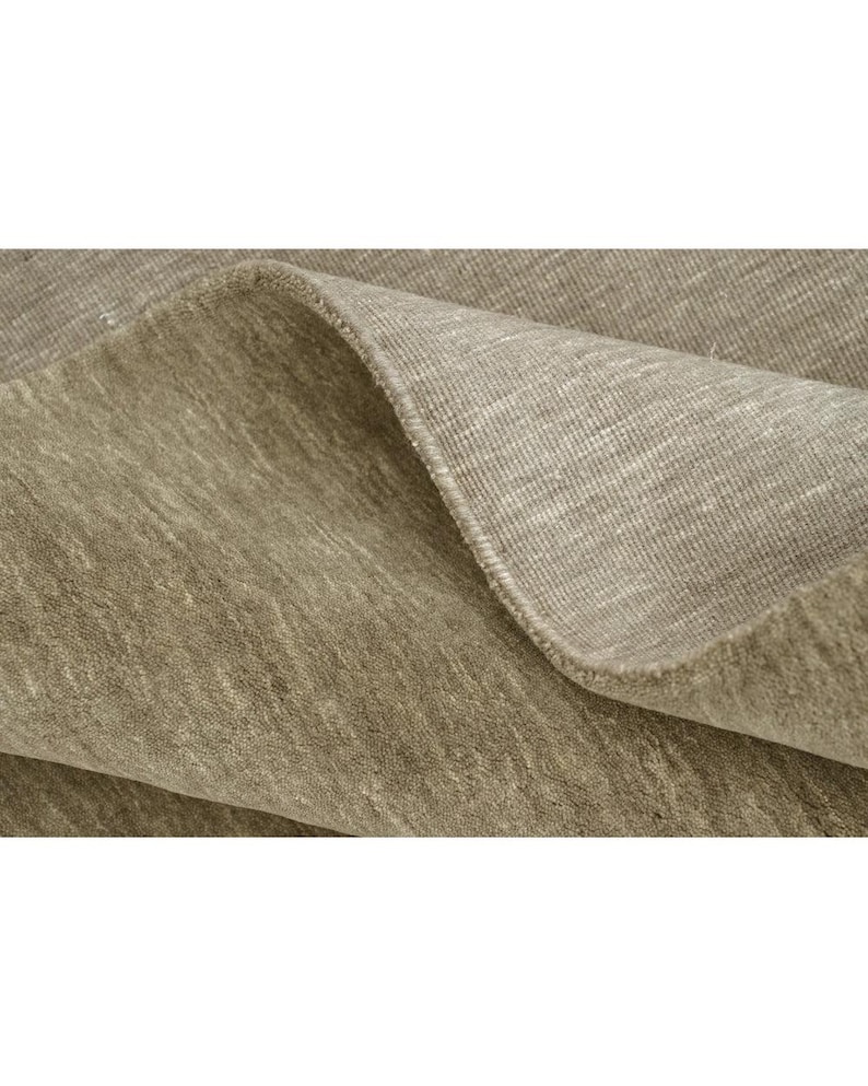 May include: A taupe-colored area rug with a dense, short-pile texture. The rug is folded over to reveal the underside, which displays a lighter, woven fabric construction with a visible grid-like pattern and a finished, reinforced edge.