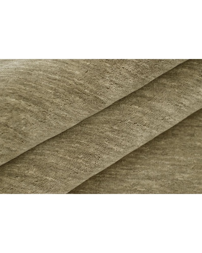 May include: A folded piece of fabric with a soft, textured surface in a muted taupe or light brown color. The material displays a dense, short pile with subtle variations in tone and light reflection across the overlapping layers of textile.