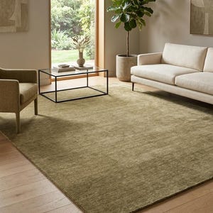 May include: A large, rectangular area rug in a solid, muted olive green tone with a low-pile, textured surface. The rug features a consistent, monochromatic color throughout, covering a significant portion of the light-colored wooden floor.